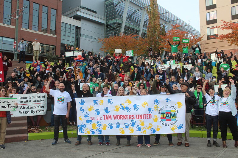 Many people stand with fists raised in the air behind a banner reading "we are TAM Workers United"