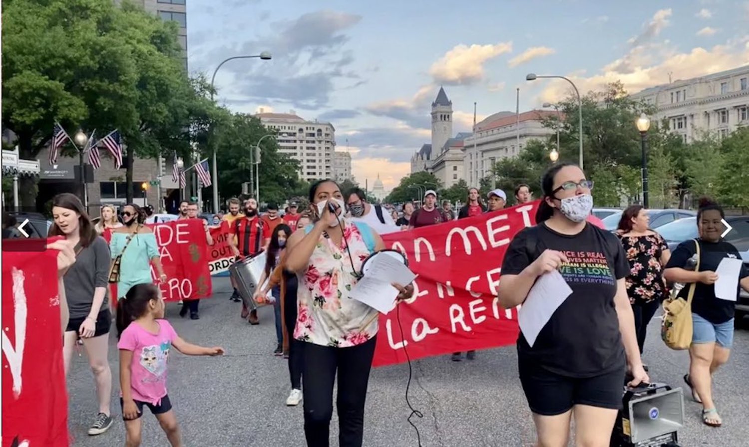A crowd of tenants and activists carrying "Cancel Rent" banners march through a street in downtown DC. Estela, a Latina tenant leader, speaks into a microphone in the center.