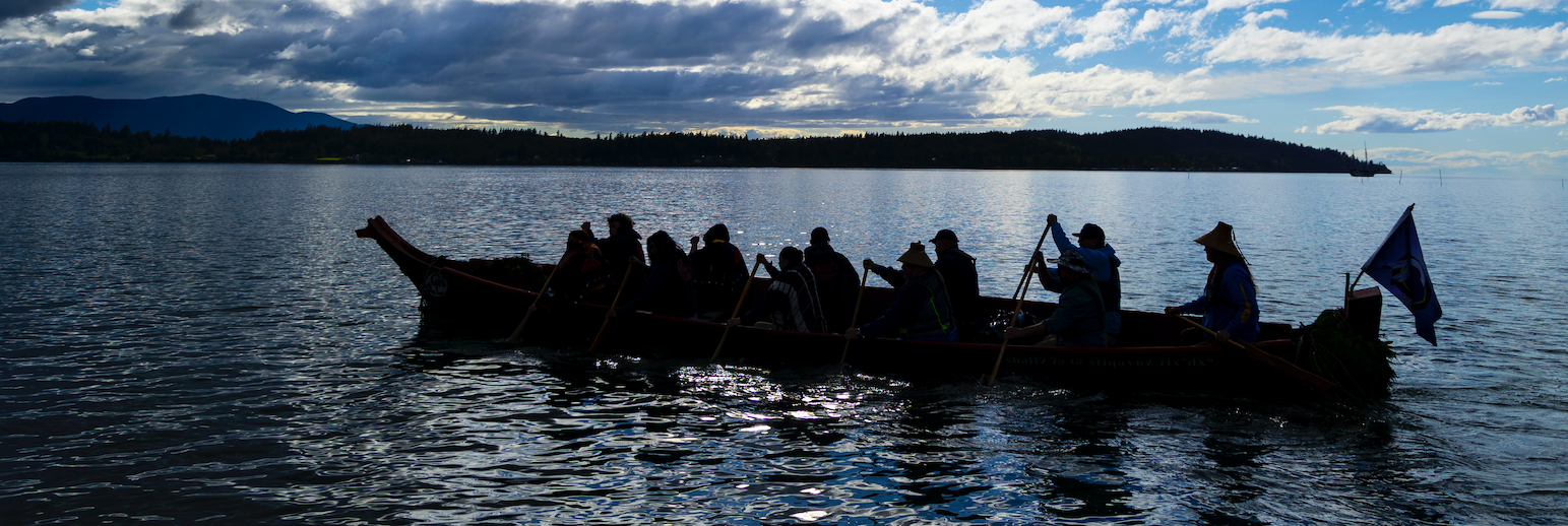 Backlit image of Indigenous paddlers in a seafaring canoe on the Salish Sea