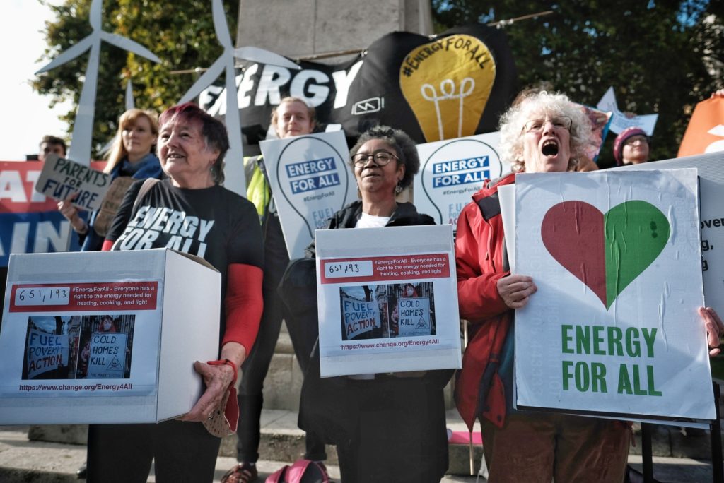 Participants at the Energy For All petition hand-in demo. Holding petition boxes with an Energy For All banner and windmill props in the background.