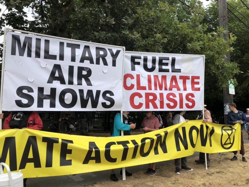 2 Large banners held by 6 people saying "Mililtary Air Shows / Fuel Climate Crisis" and "Climate Action Now"