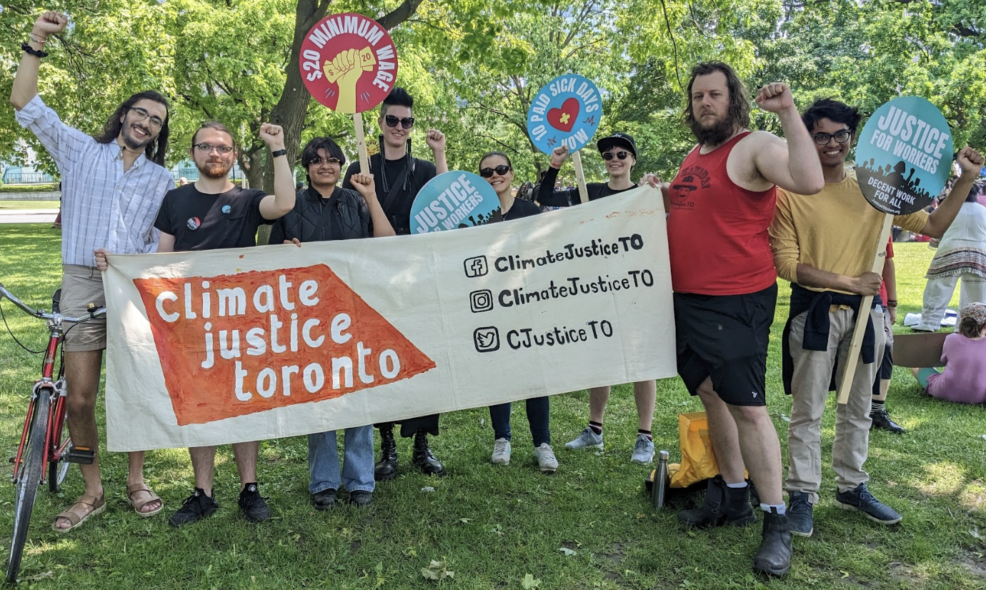 Eight people stand in a park with fists raised in solidarity. They are holding a banner that says "Climate Justice Toronto" and signs calling for justice for workers, 10 paid sick days, and a $20 minimum wage.