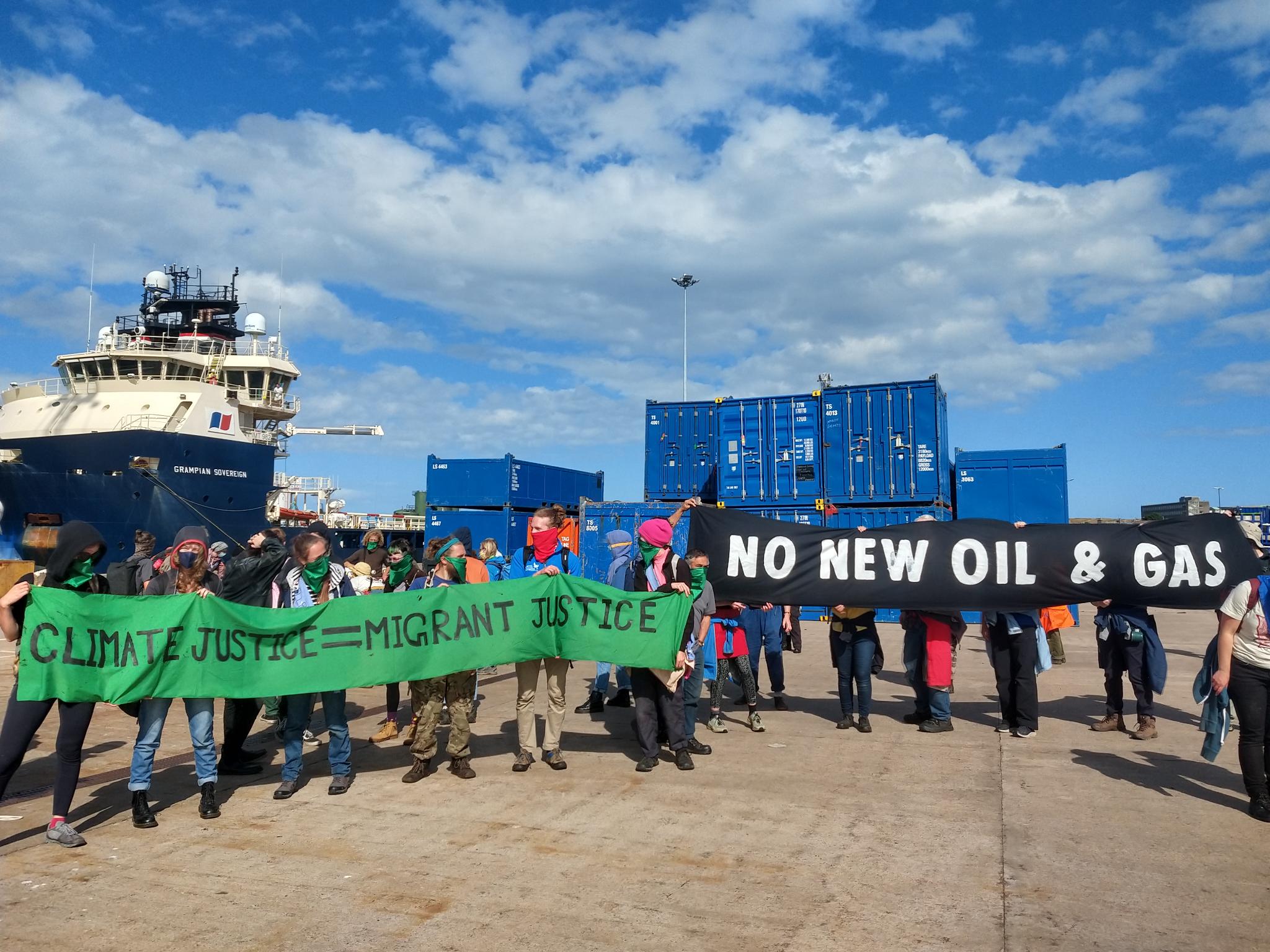 A group of around a dozen climate activists stand in front of a ship called Grampian Sovereign and crates with banners that read CLIMATE JUSTICE = MIGRANT JUSTICE and NO NEW OIL & GAS
