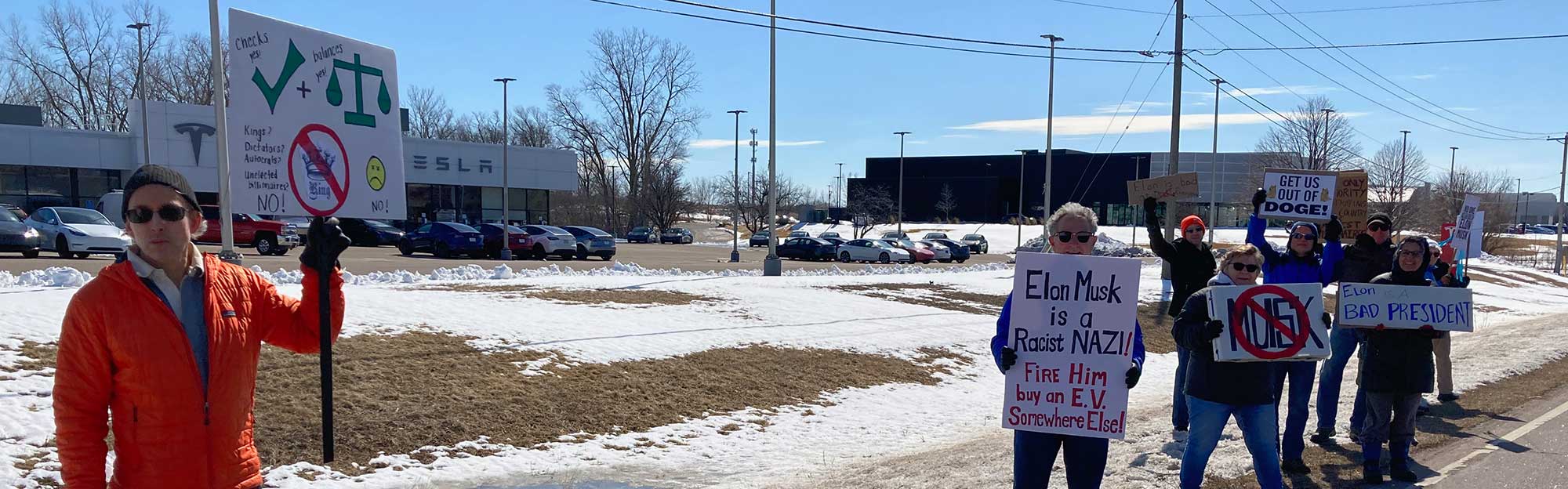 About 10 people standing with signs in front of a Tesla dealership