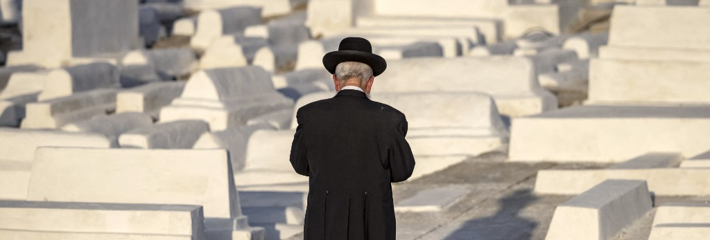 An observant Jew faces away from the camera, holding a siddur and praying over graves in a Moroccan Jewish cemetery.