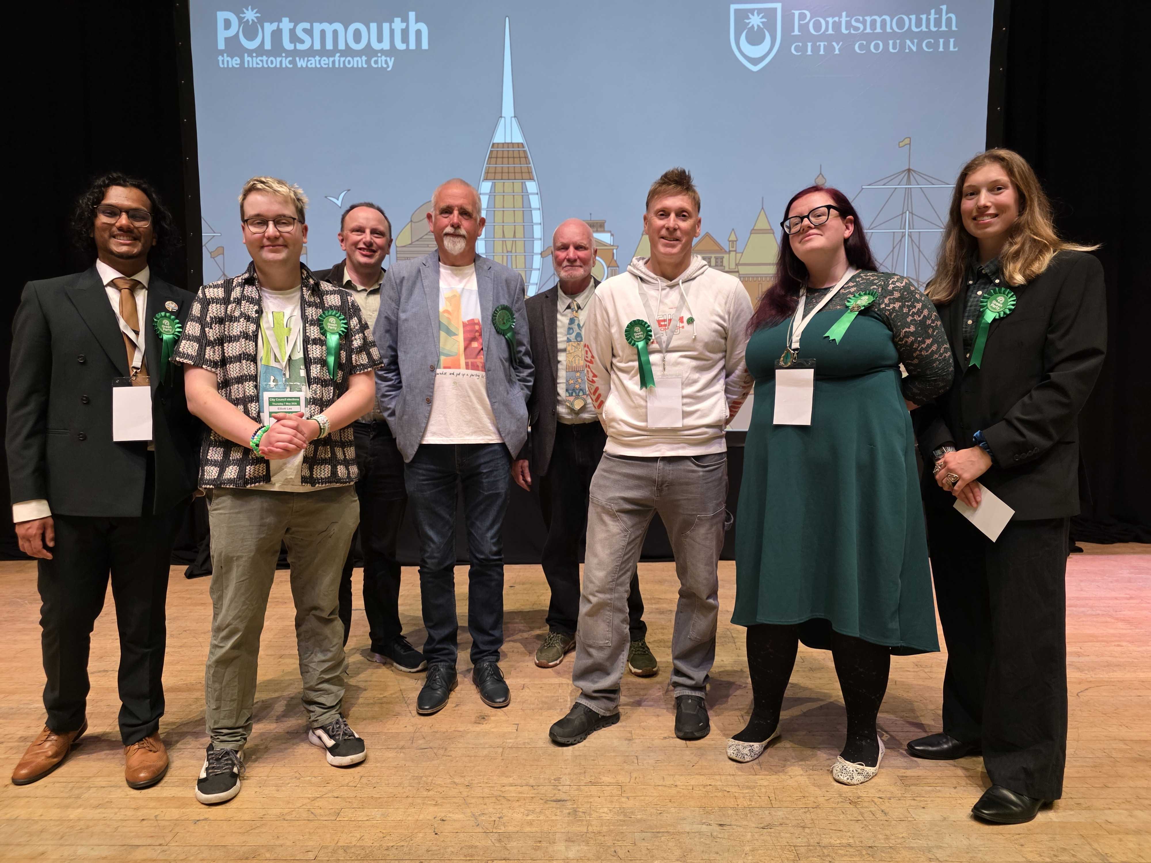 Seven Portsmouth Green Party election candidates stand together on a wooden stage in front of a projection screen displaying the Portsmouth City Council logo and Spinnaker Tower skyline. All candidates are wearing bright green election rosettes. The group includes individuals in a mix of formal suits, smart-casual jackets, and branded tops, smiling for the camera with election accreditation lanyards around their necks.