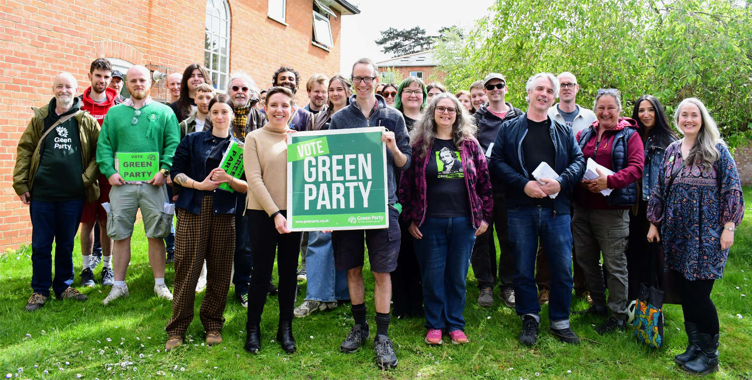 Wide shot of group of Green campaigners with candidate holding Green Party sign in centre.