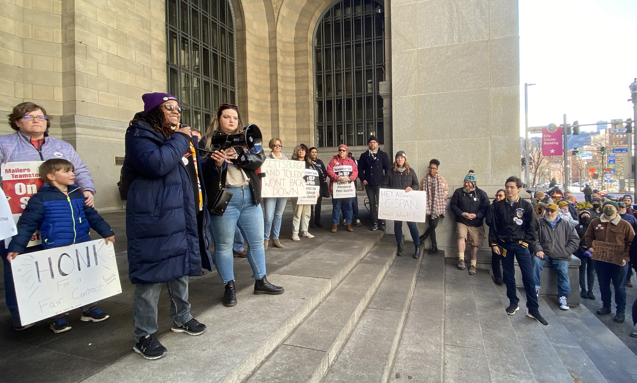 Striking workers and supporters stand outside the City-County Building in downtown Pittsburgh in January of 2023. Nila Payton of Hospital Workers Rising holds a megaphone next to Natalie Duleba, a striking editor and designer of the Pittsburgh Post-Gazette while a crowd watches in the background on a sunny day.