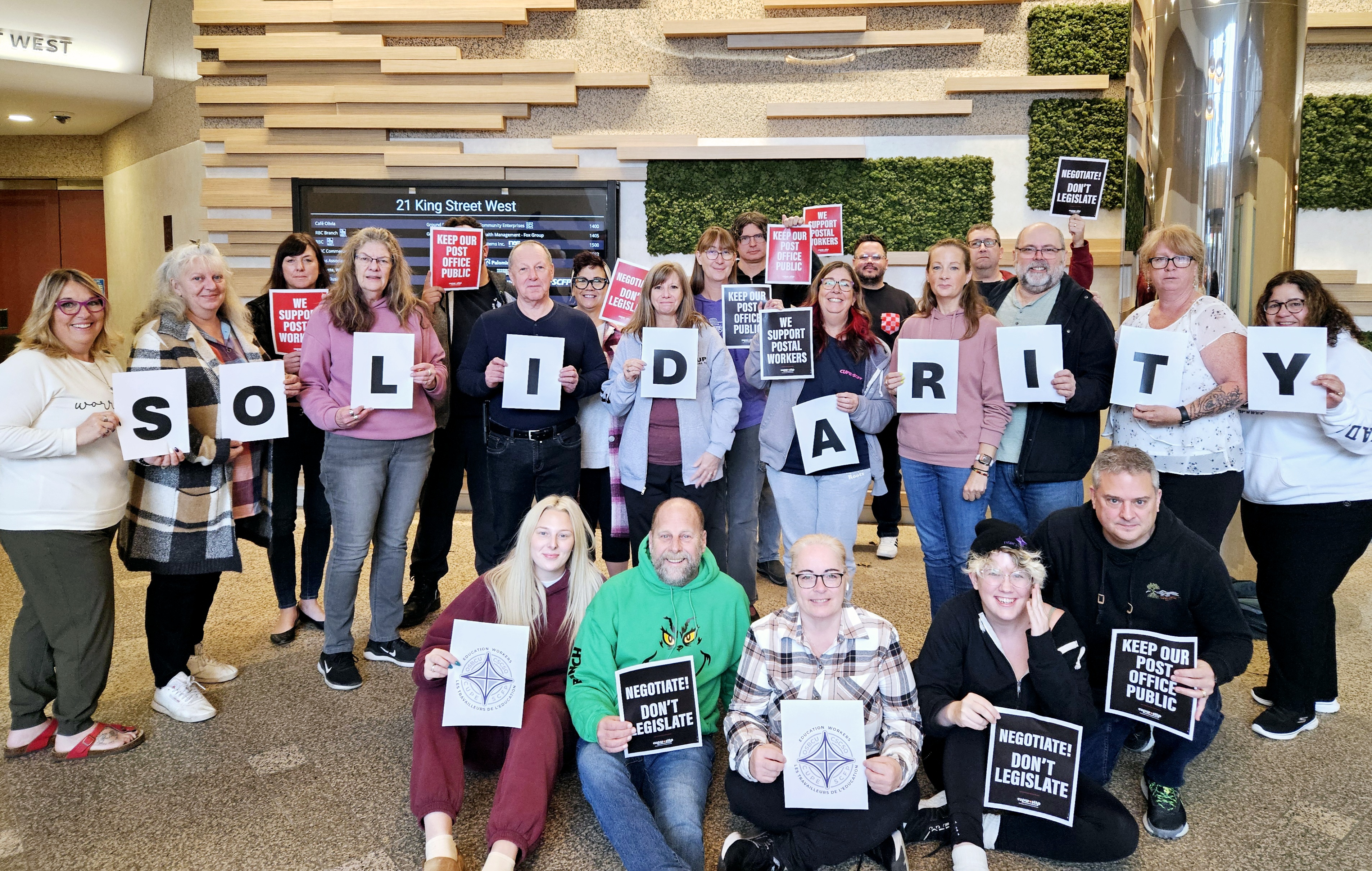 Un groupe de personnes à l'intérieur tient des lettres individuelles épelant "SOLIDARITÉ" et des pancartes avec des messages tels que "Gardez notre public public" et "Négociez, ne légiférez pas". 