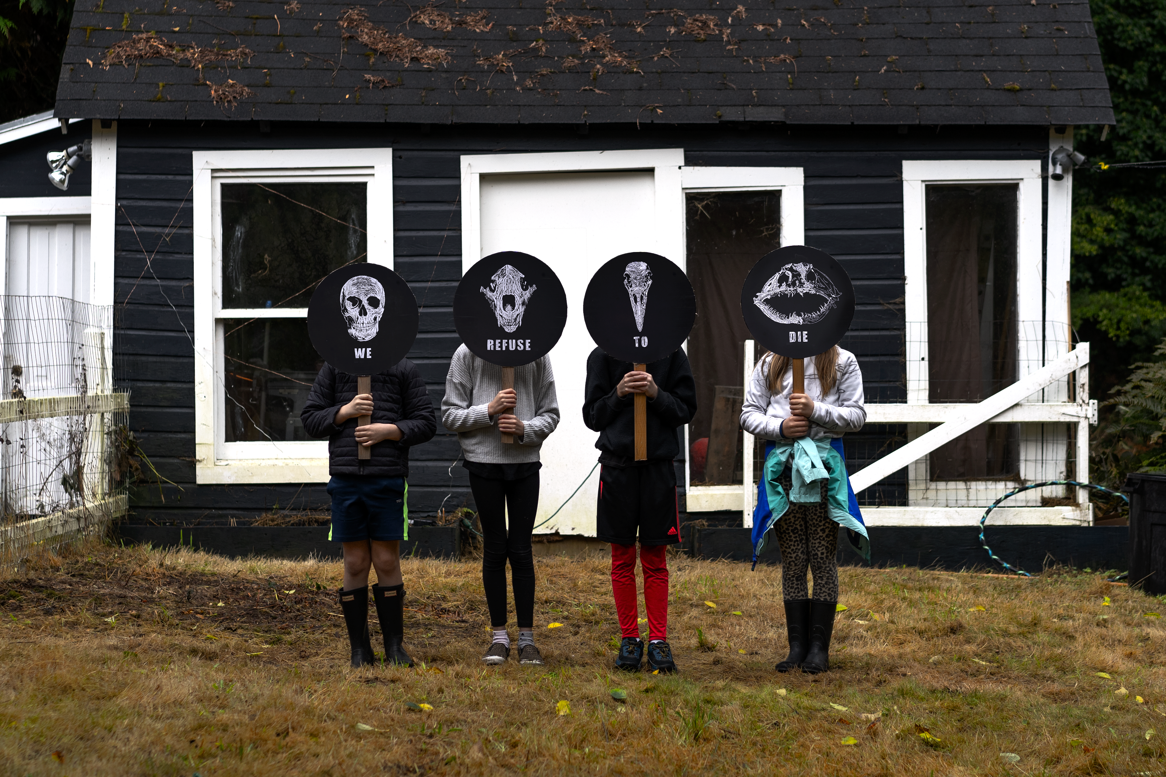 Four children stand in a row in front of a dark gray shed in the Pacific Northwest, holding circular protest signs that shield their faces from view. Printed in white on a black background, each sign illustrates a human or animal skull facing head-on, ren