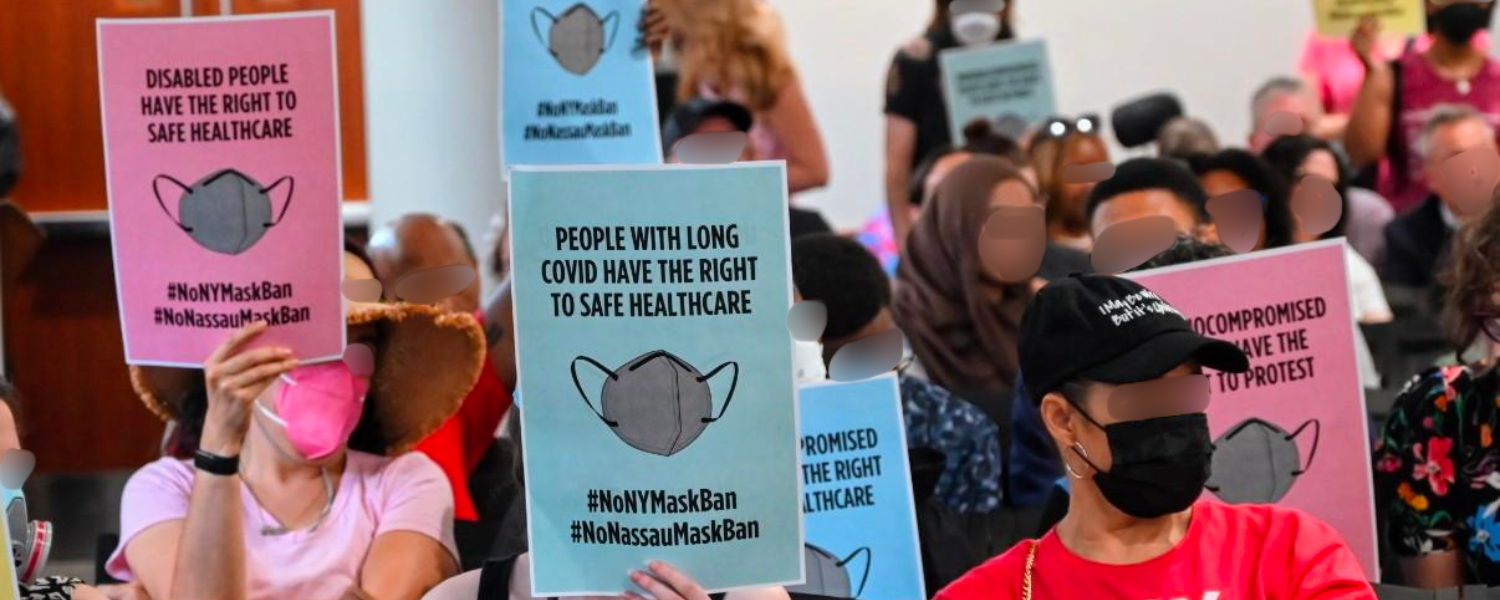 A photo of masked attendees at the Nassau County Legislature holding up colorful signs that say things like "Disabled People Have the Right to Safe Healthcare" and "People with Long COVID Have the Right to Safe Healthcare." All of the faces in the photo are blurred.