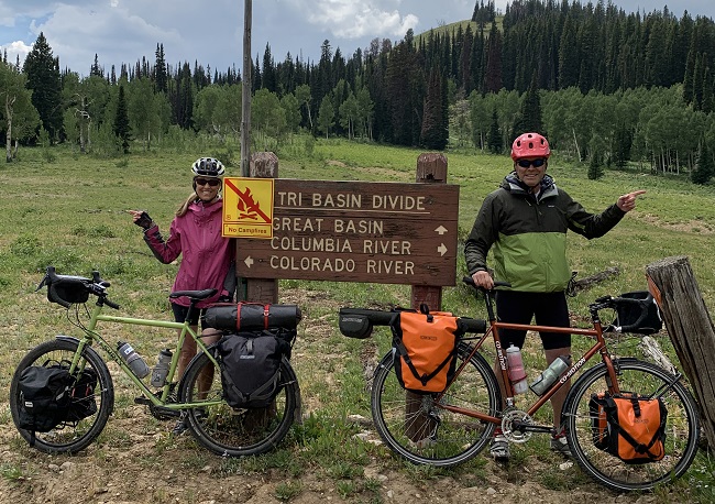 Two long distance bicyclists stand near a trail sign with two fully loaded bikes, pointed in opposite directions.