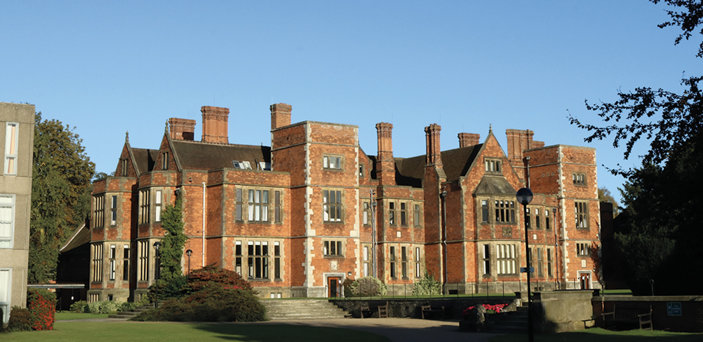 An image of Heslington Hall. Grass and trees are shown in the foreground and a blue sky in the background. The edge of a grey building can be seen on the left.