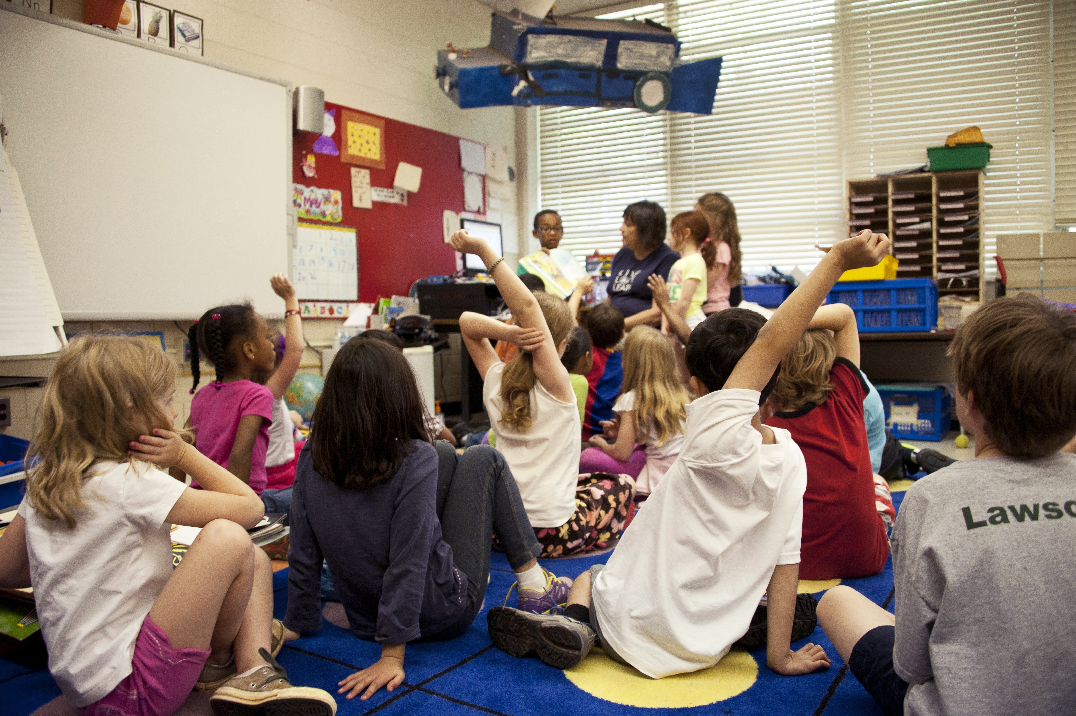 Students sitting on a rug while another student presents to the class. 