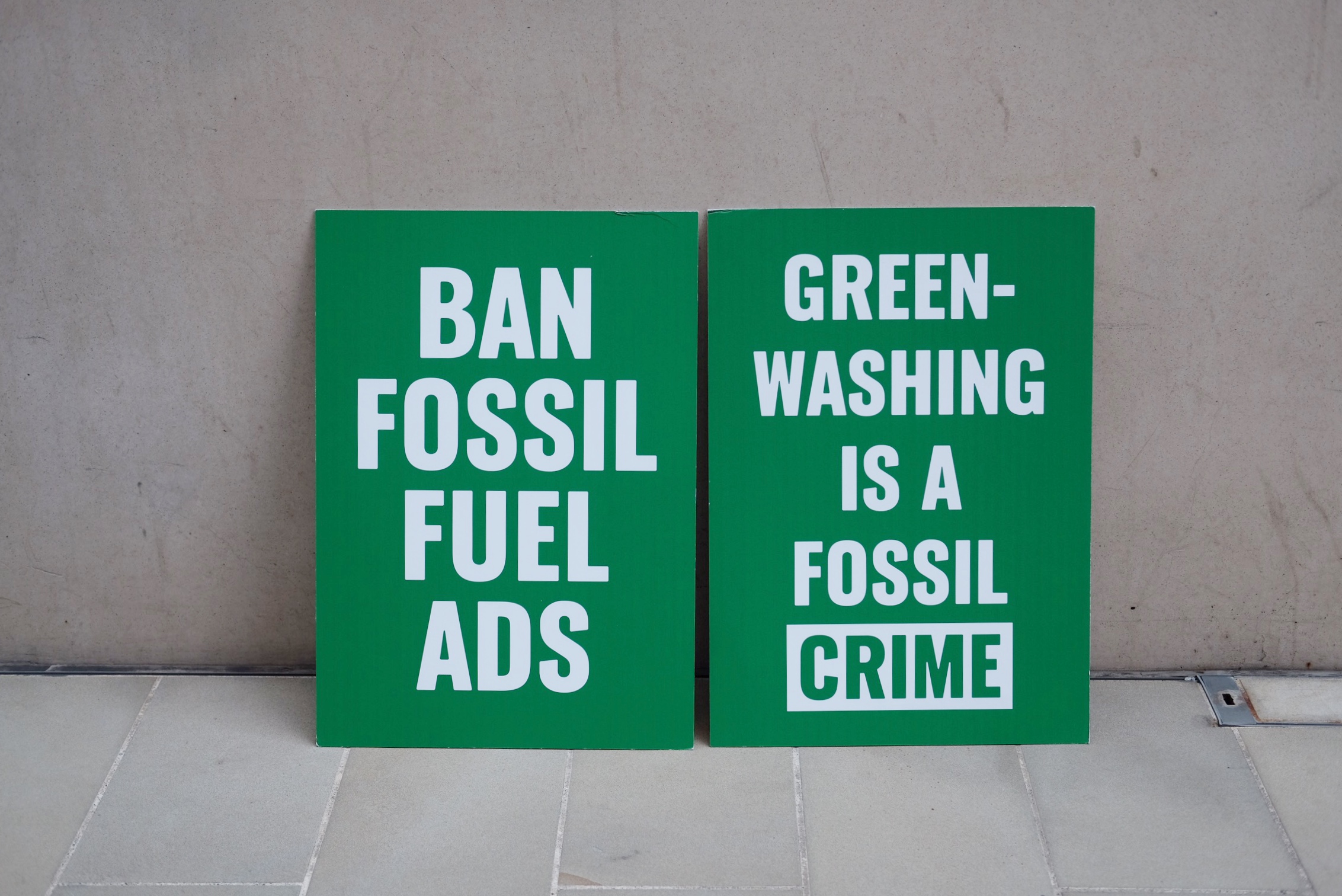 Image of two green placards leaning against a white wall with white writing in the left placard 'ban fossil fuel ads' and white writing on the left placard 'green-washing is a fossil crime'
