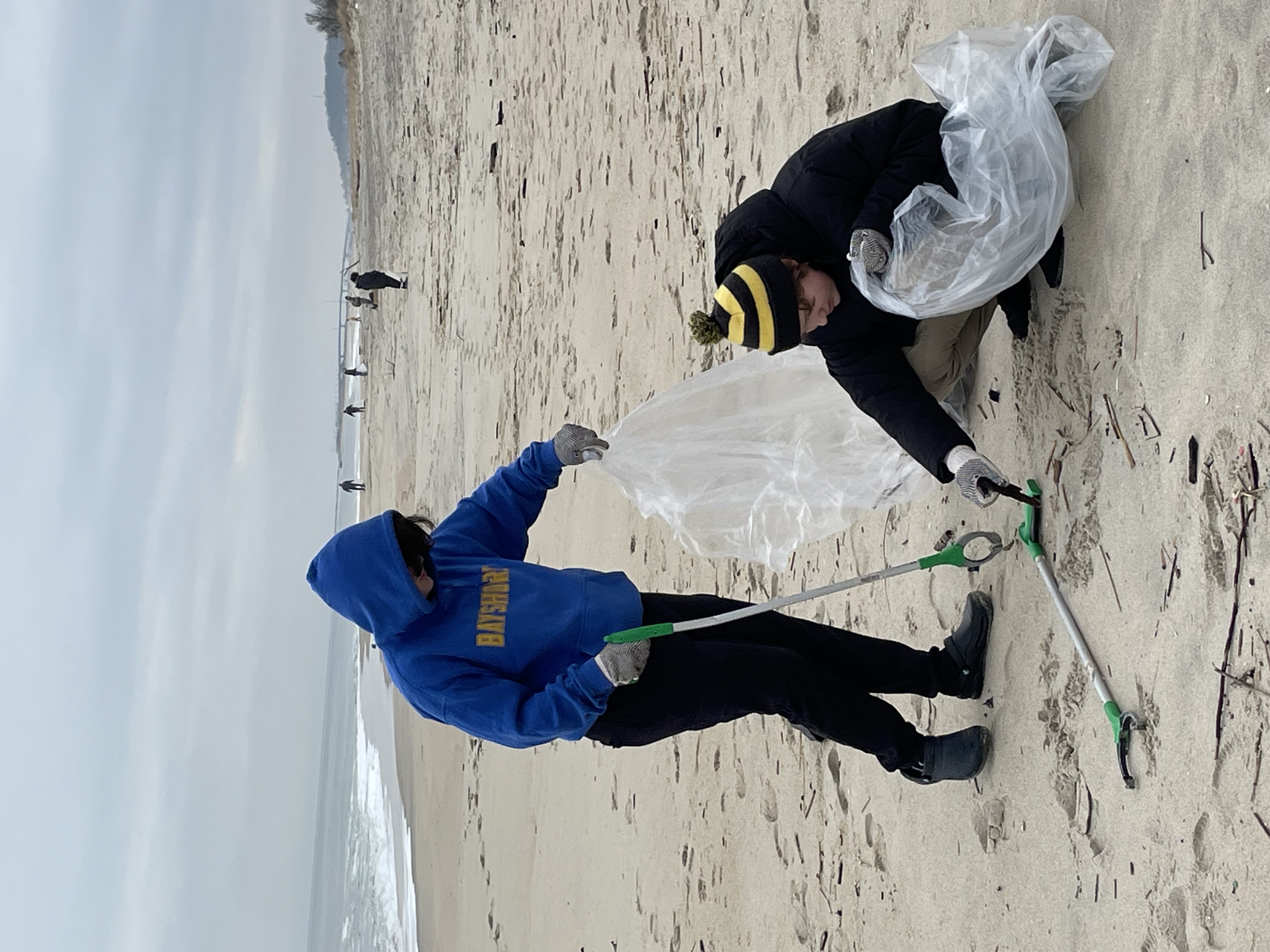 Two people wearing winter hats and gloves, helping to collect and bag waste off the beach