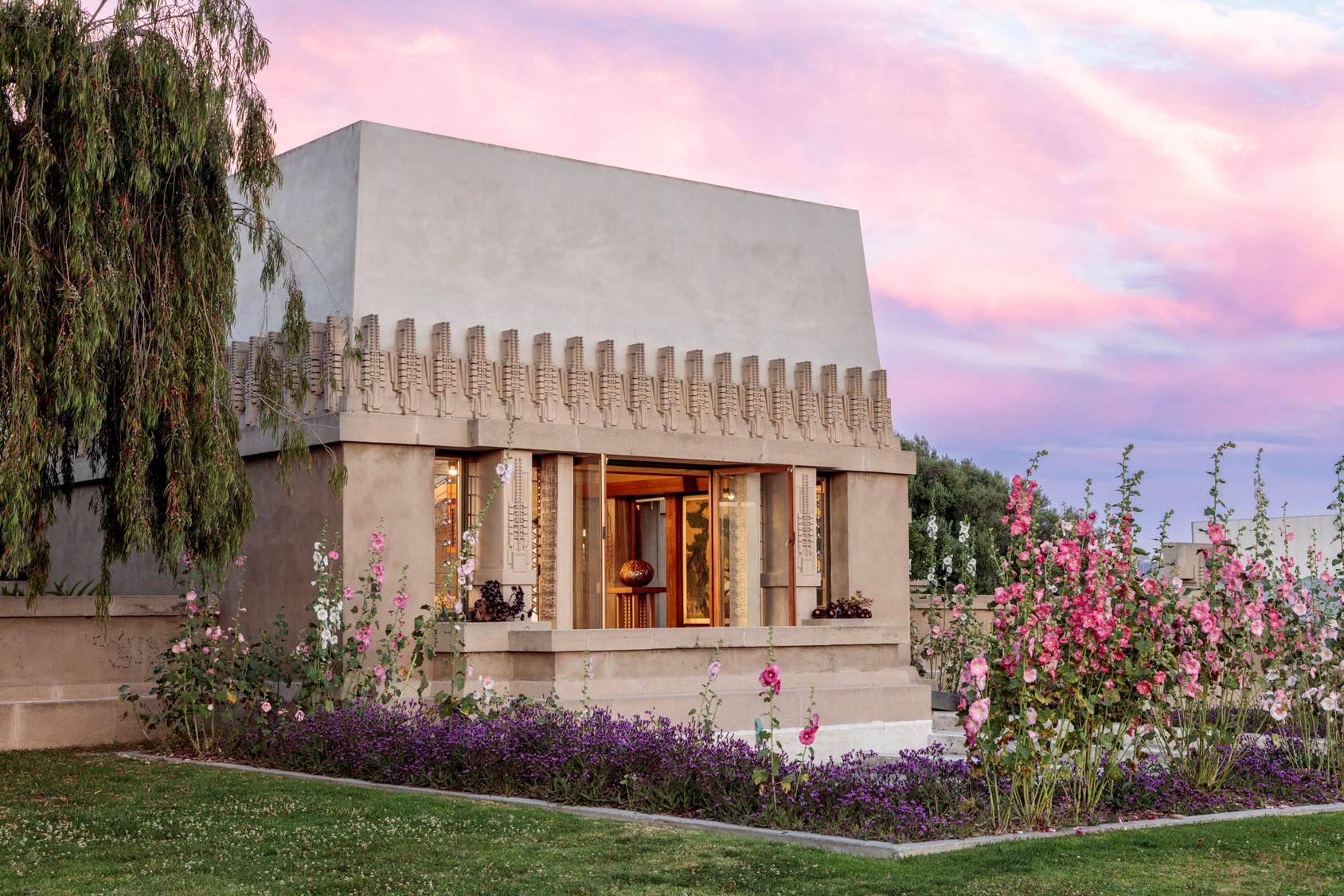 Photo of Frank Lloyd Wright's Hollyhock House in Barnsdall Art Park. House is against a pink sky with hollyhock flowers in full bloom.
