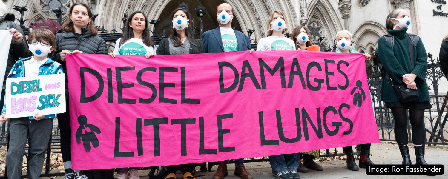 People standing with a pink banner that has the words "Diesel damages little lungs" on it in black lettering. 