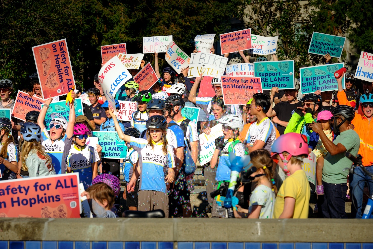 Picture of people, including children, at a rally holding signs supporting safety and bike lanes on Hopkins.