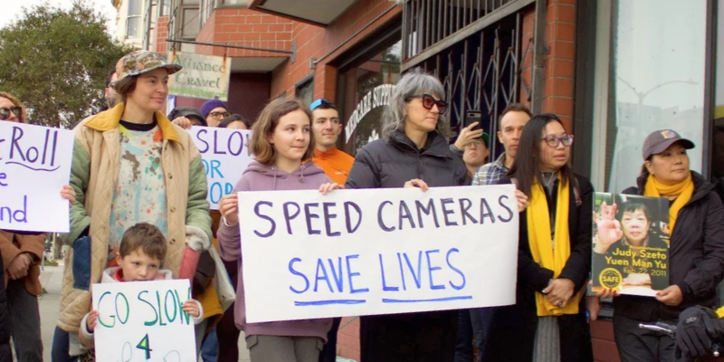 An image of two people holding a sign that says "Speed cameras save lives."