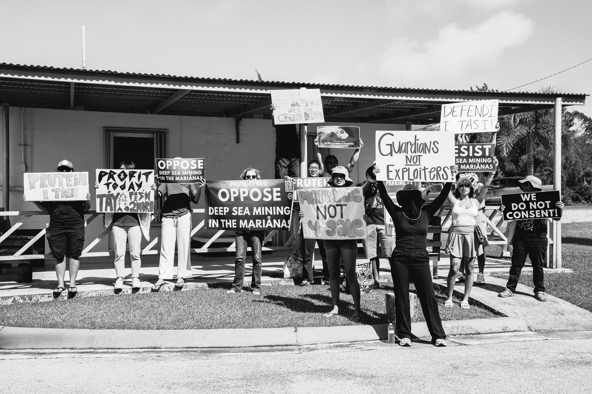 An image from a protest outside of the CNMI Office of the Governor. Banners read "OPPOSE DEEP-SEA MINING IN THE MARIANAS," "PRUTEHI I TASI," "NO DEEP SEA MINING WHEN THE OCEAN'S DYING," "DEFENDI I TASI," "PROFIT TODAY, TAYA FISH 2MORO"