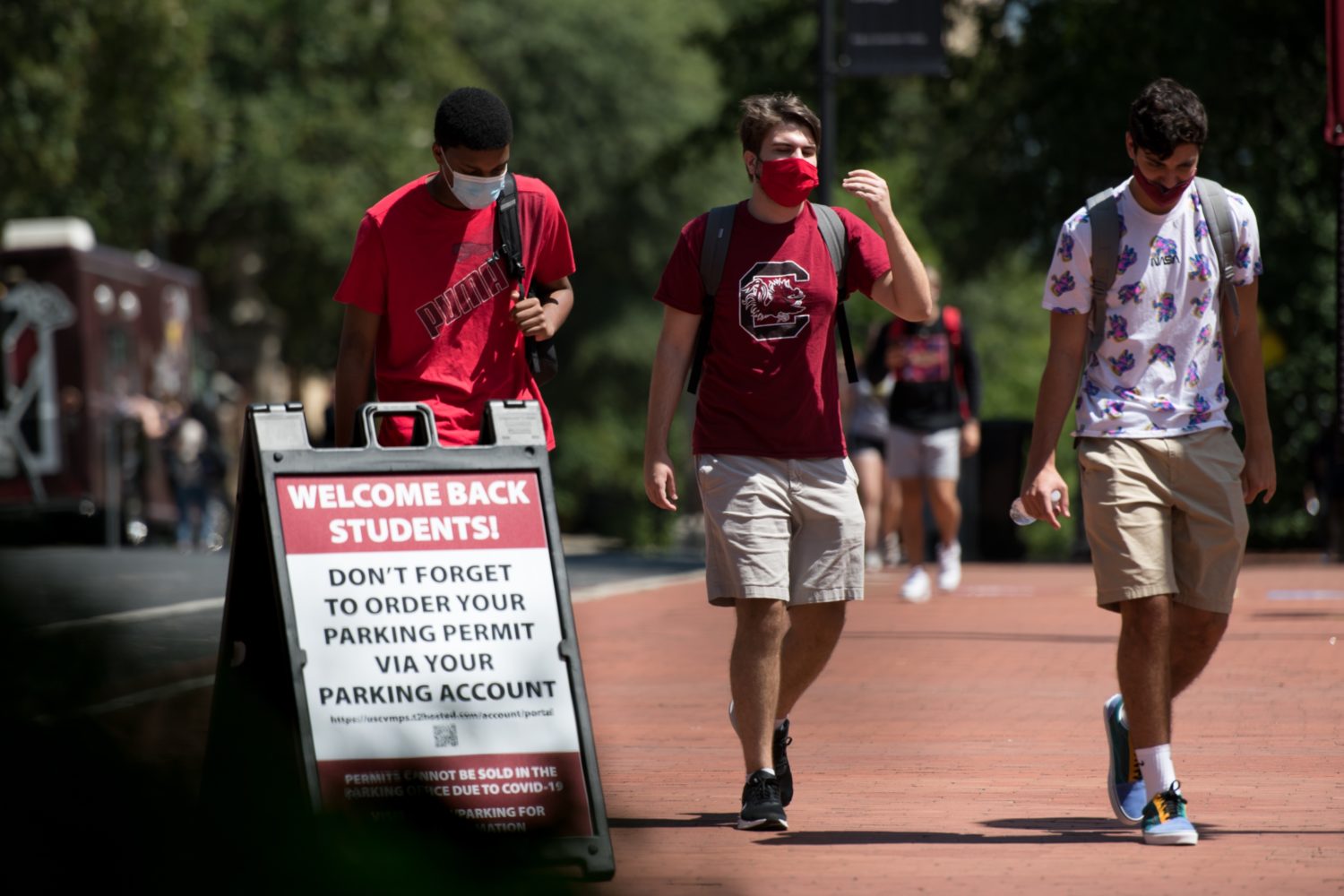 Three university students with backpacks and masks on pass outdoor sign that says "WELCOME BACK STUDENTS"