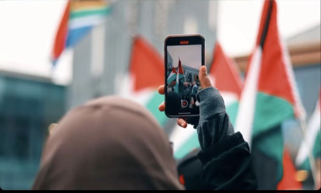 The image shows someone taking a picture at the Sheffield Solidarity Encampment. In the background is the University of Sheffield Students’ Union. There are many Palestinian flags, and a flag of South Africa.  