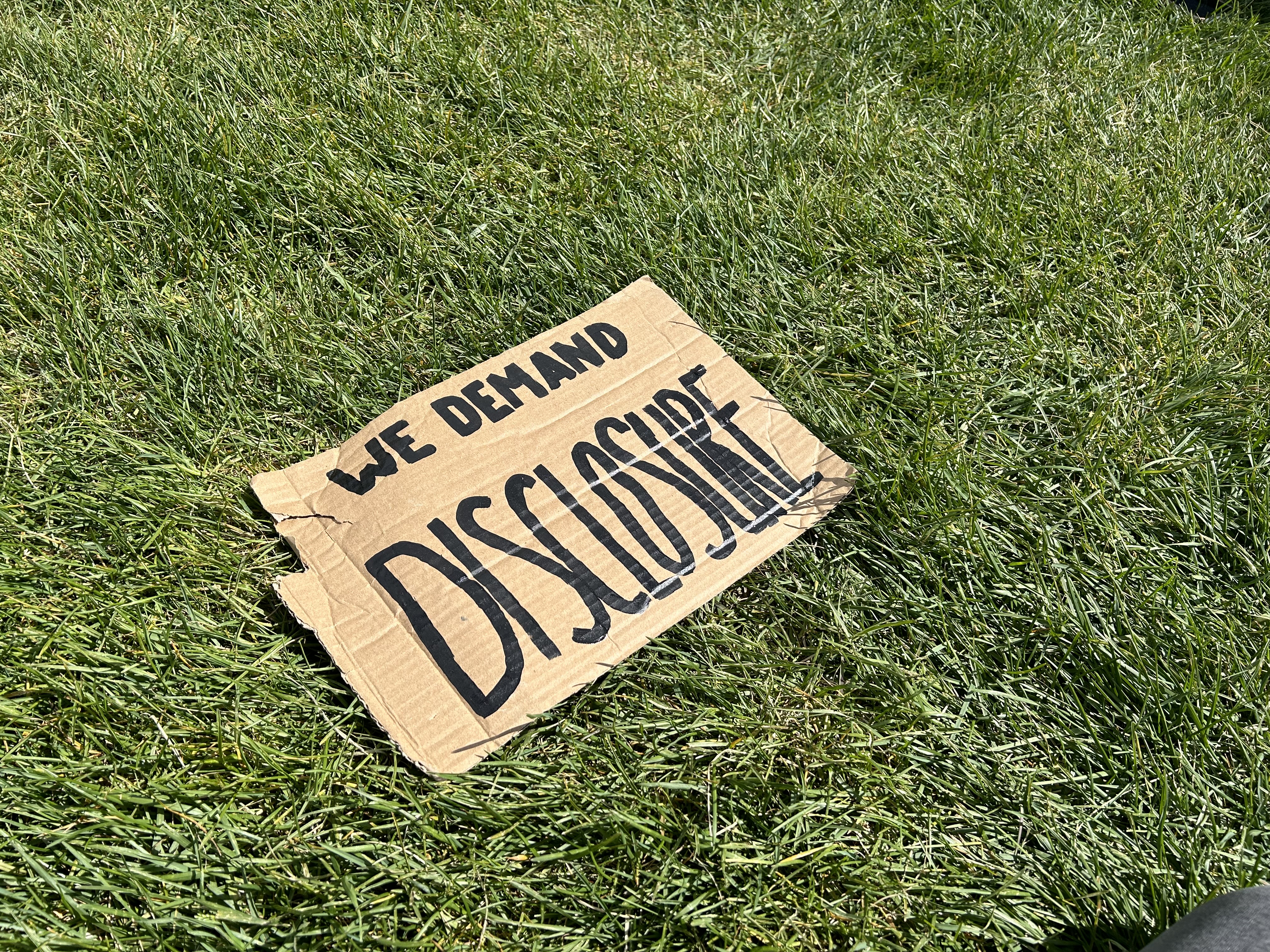 A hand-painted cardboard sign in the grass reads "We Demand Disclosure"