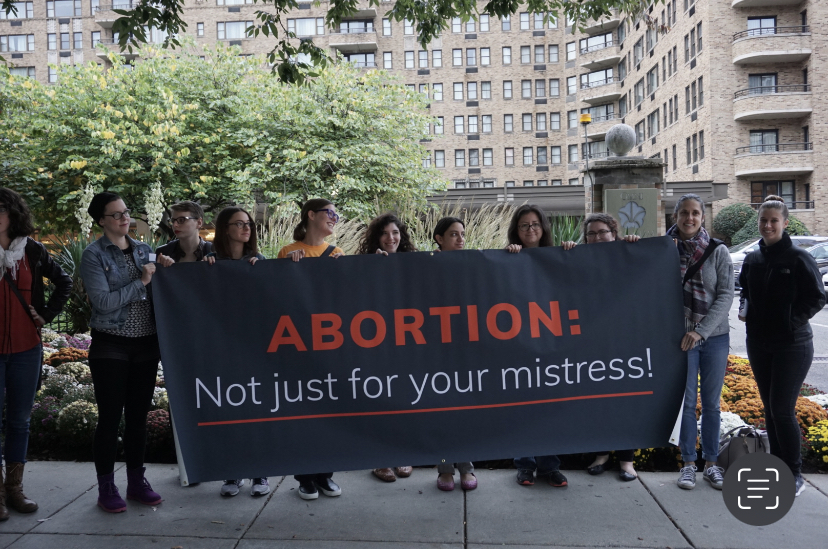 a group of people in front of a tall building holding a sign 'abortion: not just for your mistress!'