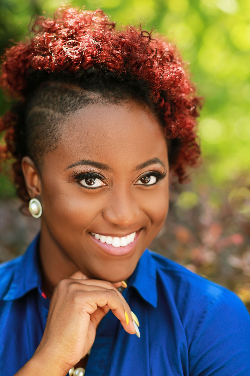 Eriqah has red hair with a side buzzcut. She is wearing big round earrings and a royal blue button up shirt. She has a big bright smile on her face.