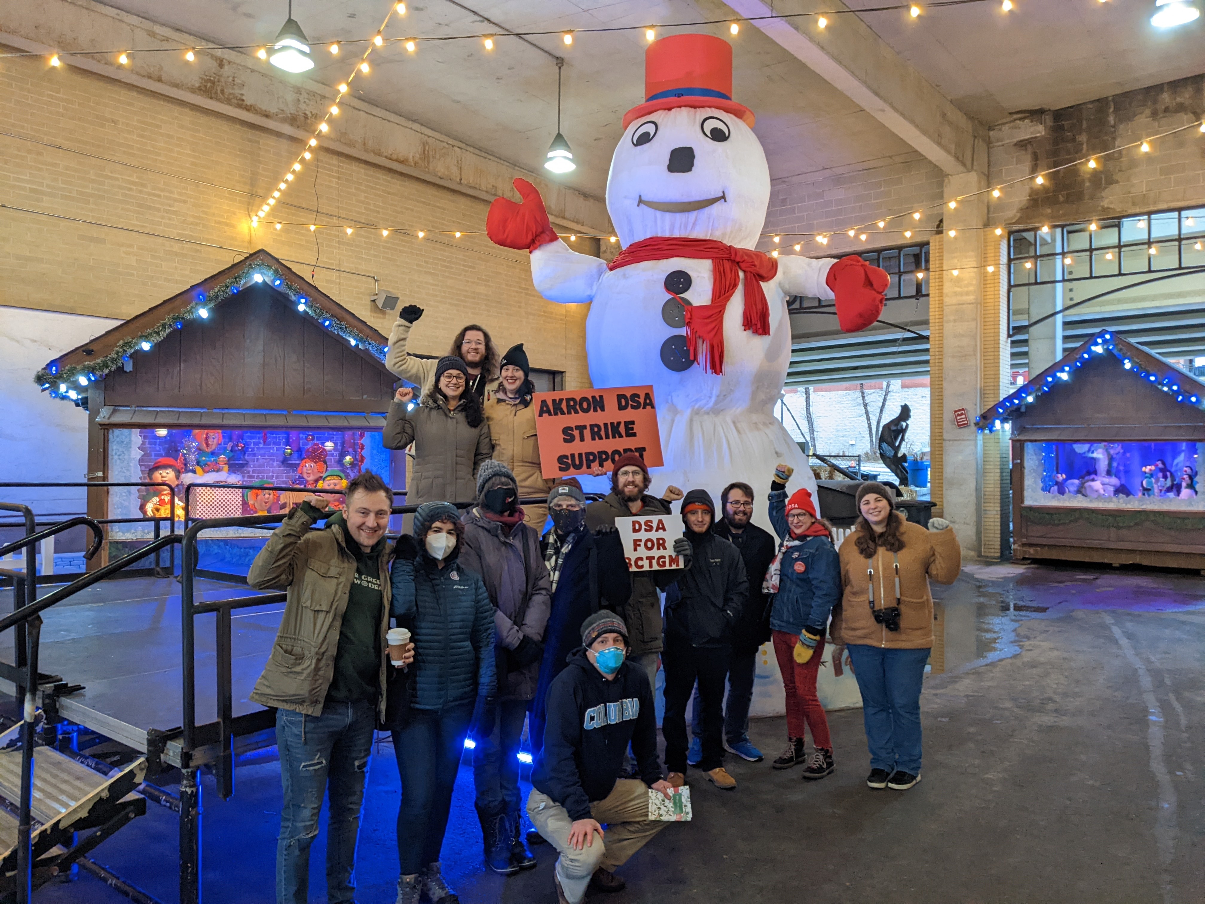 Members of Akron DSA wearing winter clothing stand in front of Archie the snowman holding a sign that says "Akron DSA Strike Support" in 2021