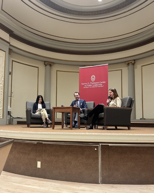 Two women sit on opposite ends of a stage with a male moderator in between. 