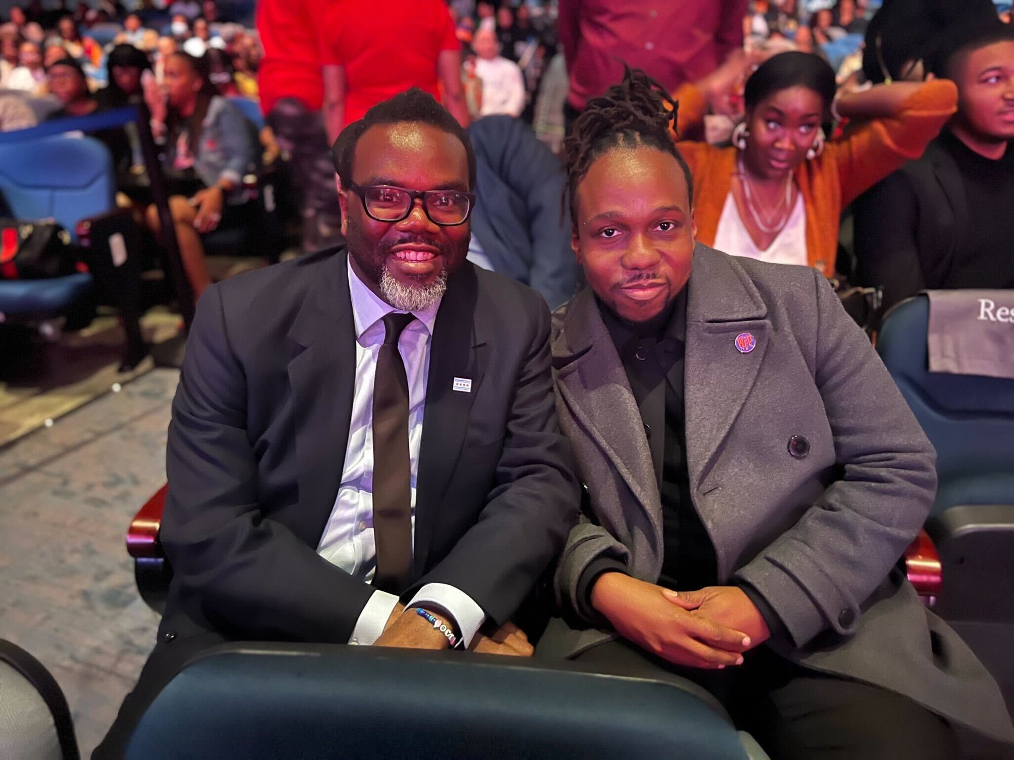 Brandon Johnson and Maurice Mitchell posing for a photo and smiling. Both Brandon and Maurice are stitting down in the audience of a big convention hall with a crowd behind them.