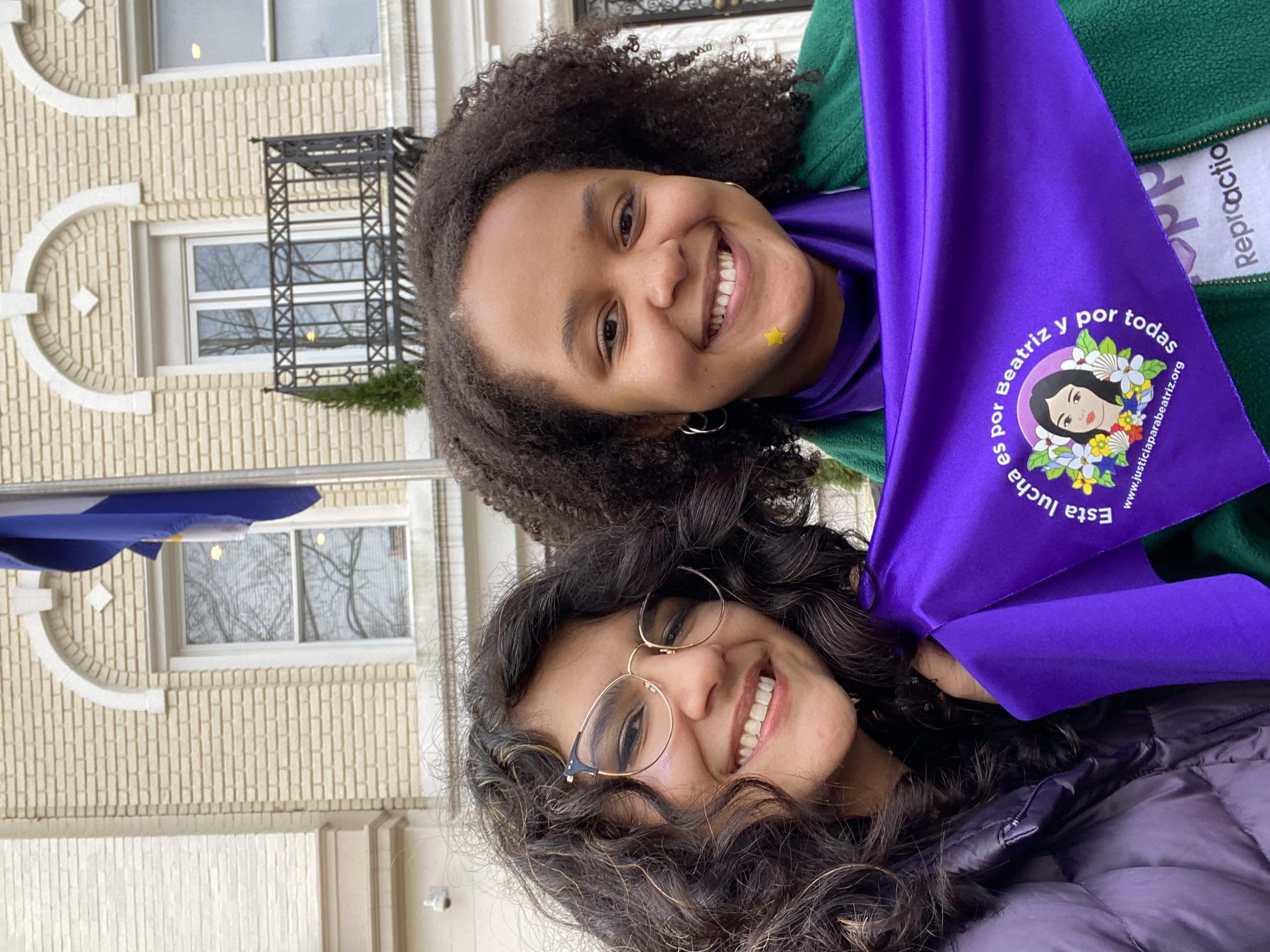 [alt image text: two people smiling, holding a purple bandana reading 'esta lucha es por beatriz y por toddas' in front of a light brick building.