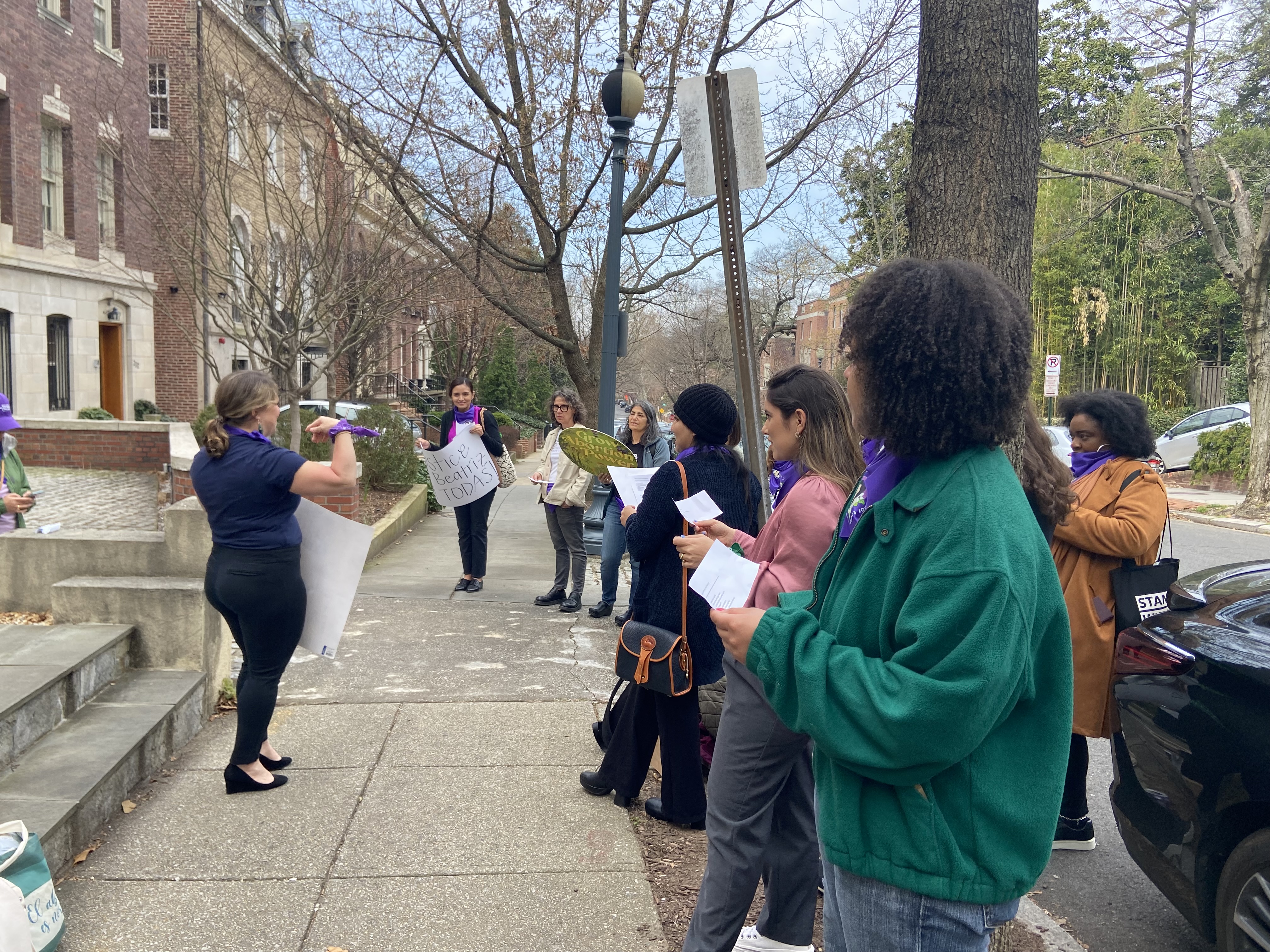   [alt image text: a group of people holding signs and papers chanting in front of a sidewalk, with one person in the center leading chants with a big sign]