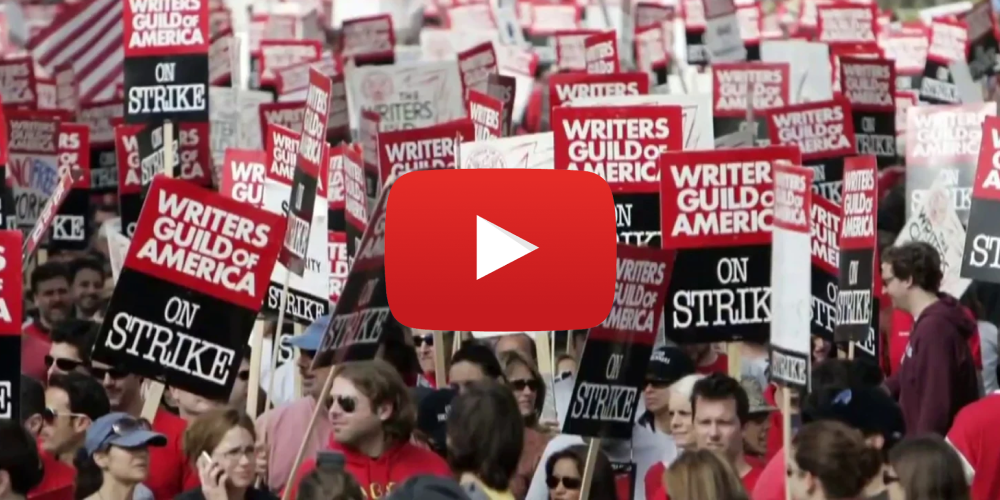 Screencap of video with people at a mass protest holding signs that say "Writers Guild of America on Strike"