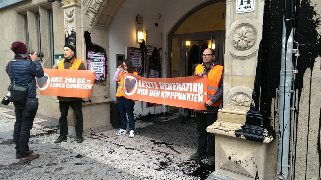 Last Generation activists hold banners outside a building smeared with black paint