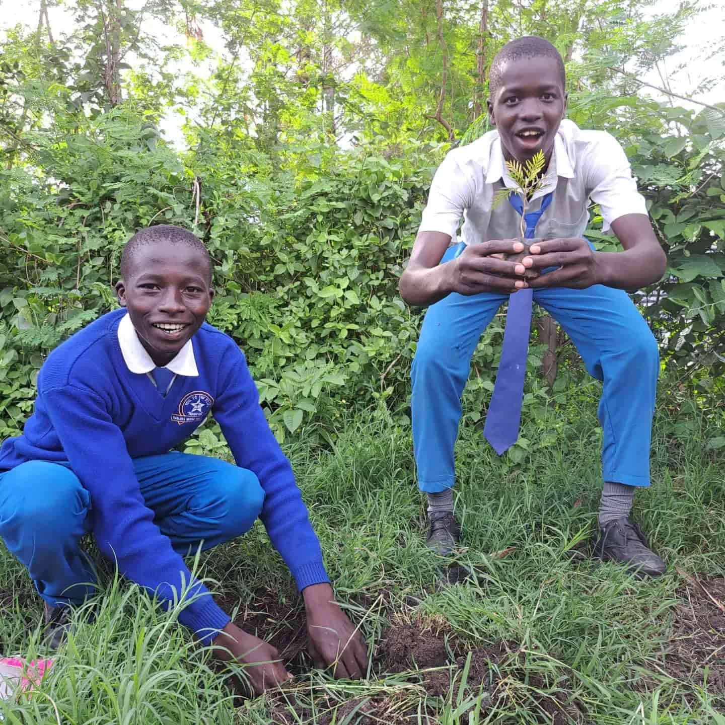 Two school boys smile, one holding a small tree sapling about to be planted