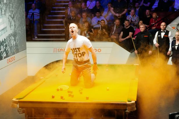 A Just Stop Oil activist shouts among a blume of orange dust, knelt on a snooker table