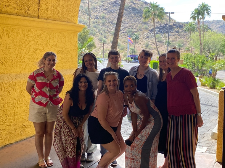 Reproaction staff gather in a group and smile at the camera in front of desert palm trees. 