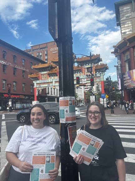 Two people stand in front of a street sign that has a self-managed abortion flyer taped to it. They are holding stacks of the same flyer. 