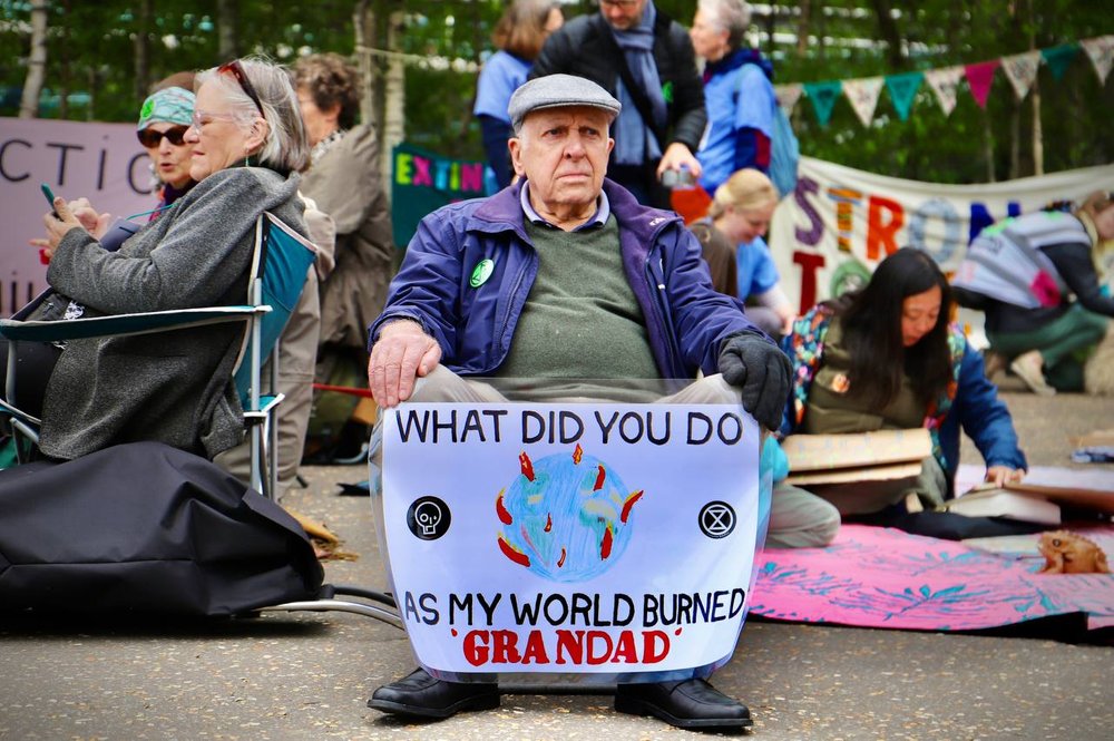 A solemn old man sits with a sign that says: What did you do as my world burned grandad?