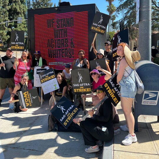 Photo of mobile billboard saying "We stand with actors and writers! Working Families Party" with protestors holding signs saying "SAG-AFTRA on strike!"