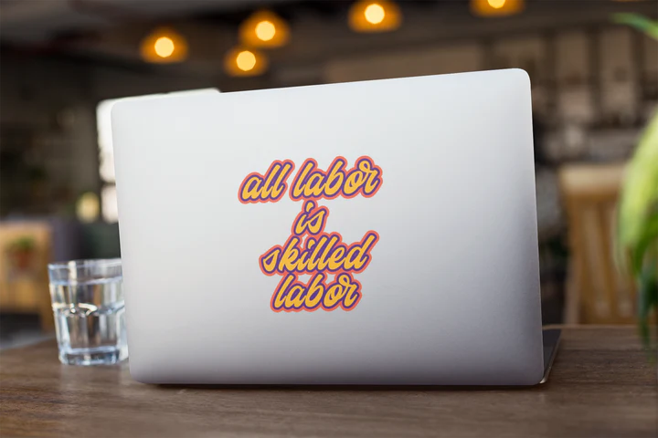 A silver laptop with a sticker on the back with orange lettering that reads all labor is skilled labor