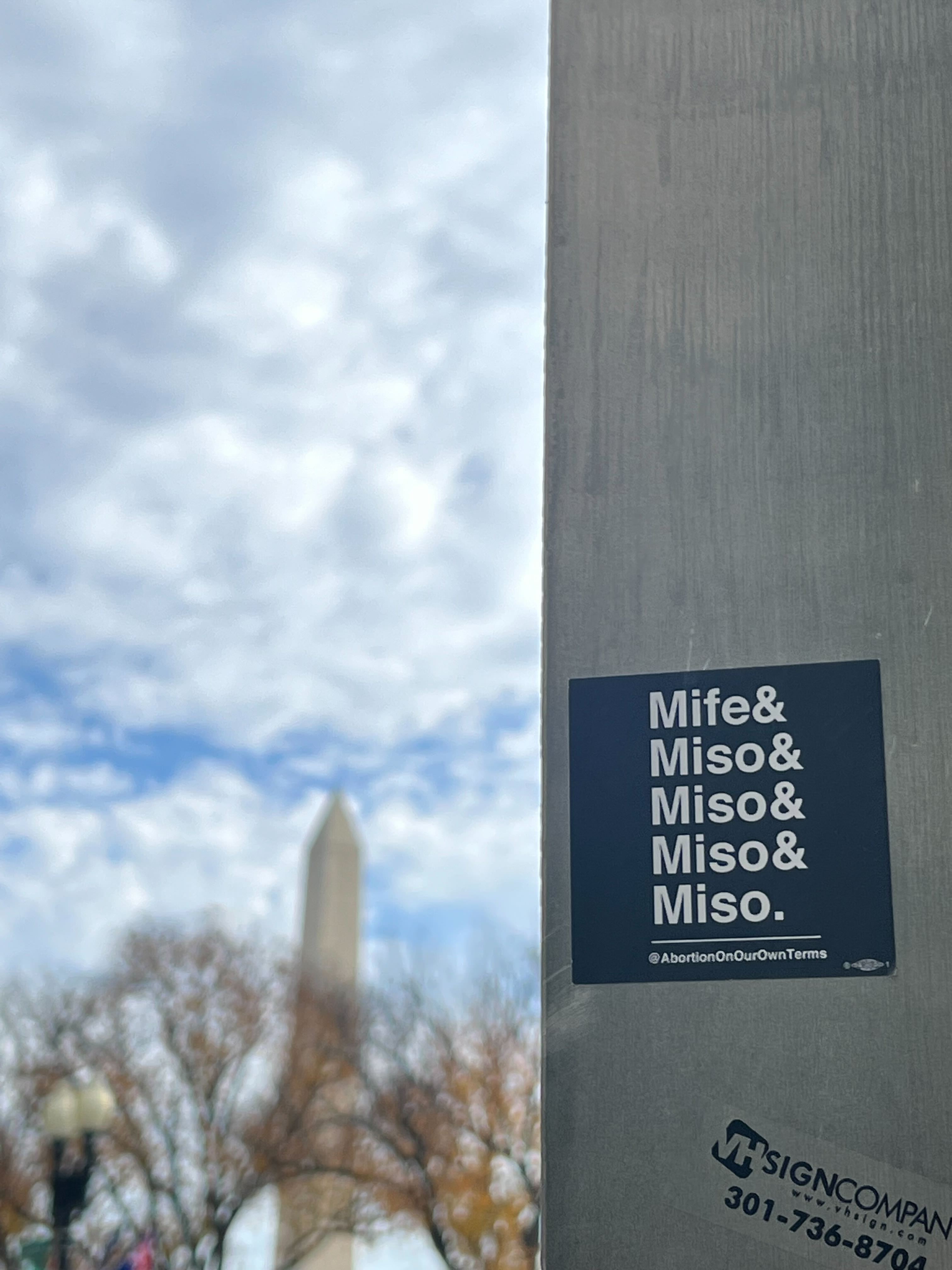 A picture of two signs saying "Mife & Miso & Miso & Miso & Miso" attached to an electrical box. The backdrop is a busy street and the Washington Monument.