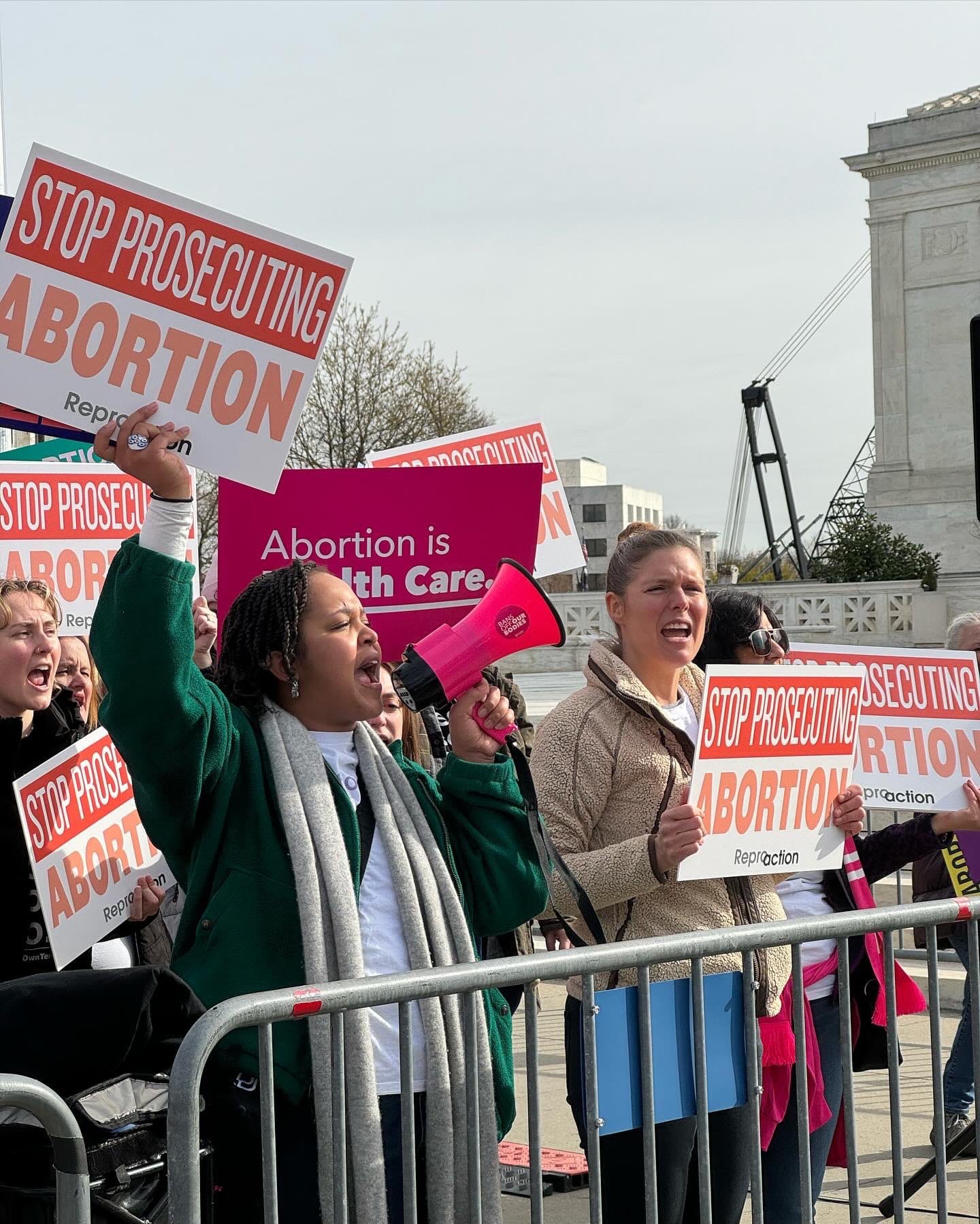 Two people stand in a crowd behind a police barrier. One yells into a megaphone while the other chants. The crowd holds signs that read Stop Prosecuting Abortion and Abortion is Health Care.