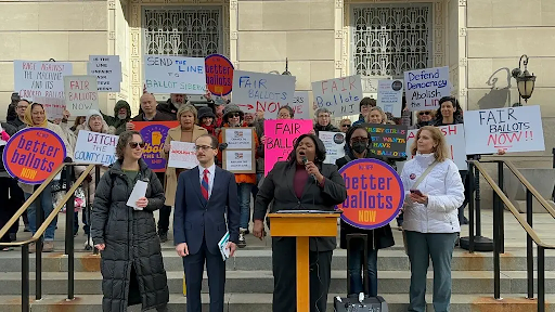 Photo of a protest against hte ballot line system with NJWFP State Director Antoinette Miles center speaking at a podium. Protestors hold signs behind reading "Better Ballots Now," "Ditch the County Line," "Defend Democracy, Abolish the Line," and more.