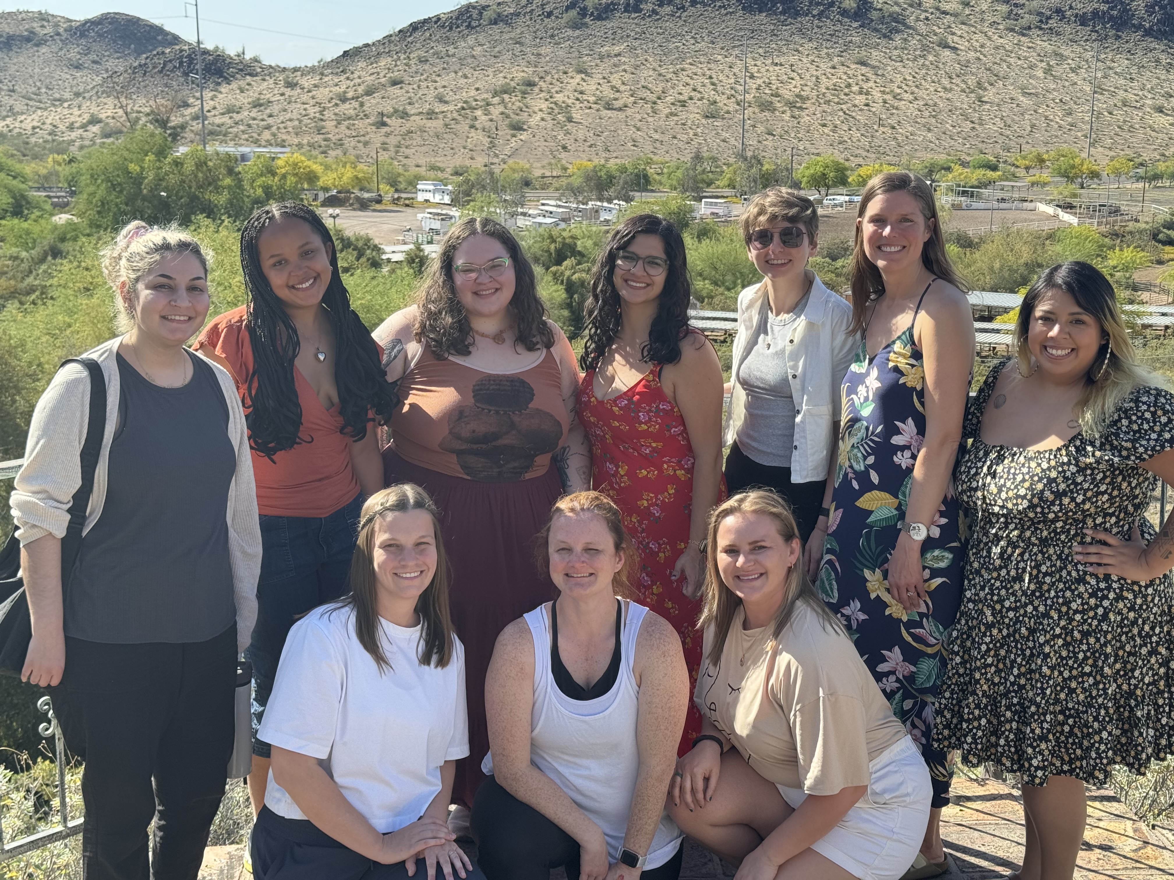 Alt-text: A group of people in dresses, shorts, pants, and shirts, arranged in two rows, the back standing, the front kneeling, in front of a desert mountain vista.