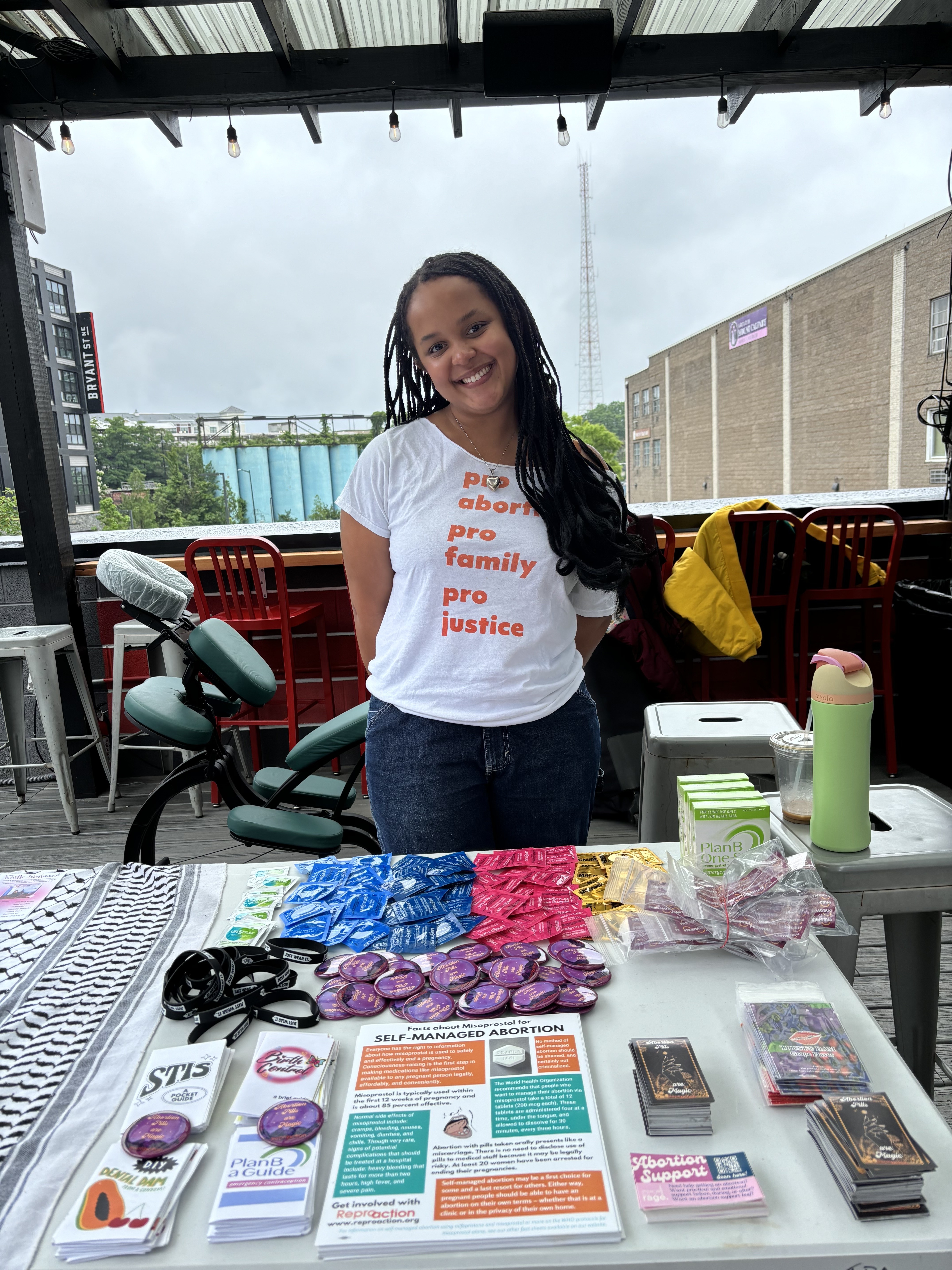 A person wearing a white shirt reading “pro-abortion pro-family pro-justice” stands in front of a table filled with outreach items such as flyers, buttons, pamphlets, and stickers.