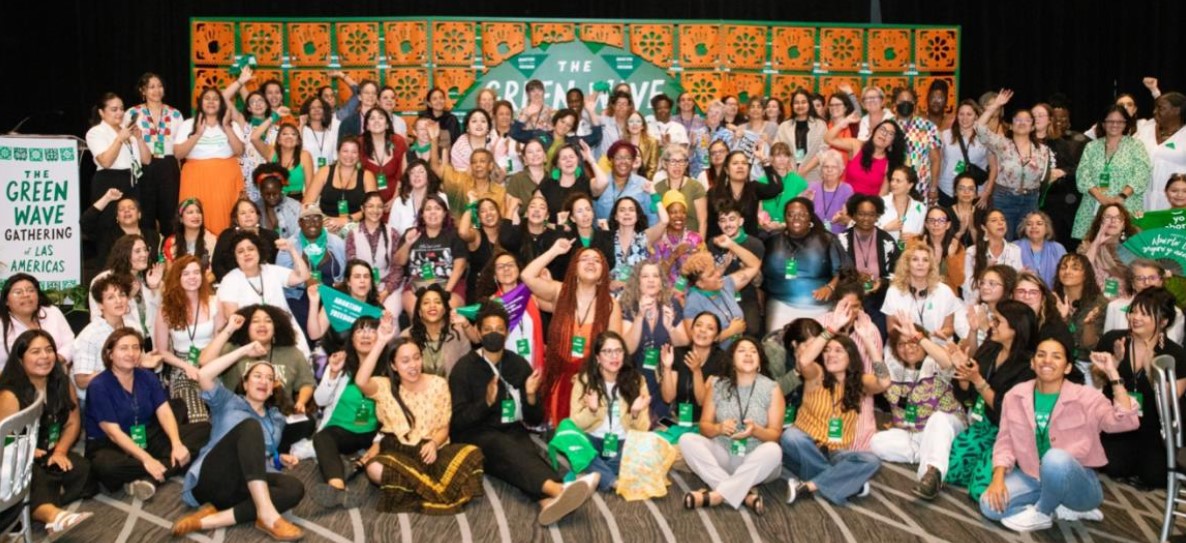 t: A group of dozens of people stand or sit, some with raised fists, in front of a banner image. A sign to the side reads: The Green Wave Gathering. 